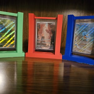 Könnte beinhalten: Drei farbenfrohe Karten-Ausstellungsrahmen in Grün, Rot und Blau. Jeder Rahmen hält eine Sammelkarte, mit der Bulbasaur-Karte im grünen Rahmen, einer Charizard-Karte im roten Rahmen und einer Squirtle-Karte im blauen Rahmen. Die Rahmen stehen auf einer Holzoberfläche.