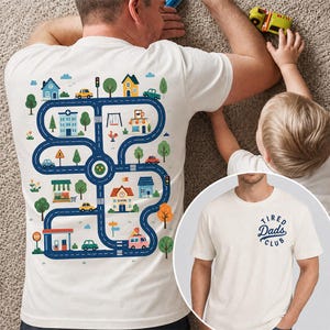 Könnte beinhalten: Cremefarbenes T-Shirt mit buntem Straßenkarten-Design auf dem Rücken, mit Häusern, Bäumen, Autos und einer Tankstelle. Auf der Vorderseite des Shirts steht in Marineblau der Text "Tired Dads Club". Ein Kind spielt in der Nähe mit einem Spielzeugauto.