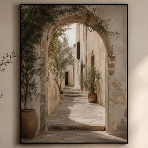 Può includere: Una stampa incorniciata raffigura un vicolo soleggiato con porte ad arco e muri in pietra. Ulivi in vasi di terracotta costeggiano il sentiero, guidando lo sguardo verso la luce lontana. L'opera d'arte è esposta sopra un mobile in legno, con una poltrona beige e un tavolino rotondo.
