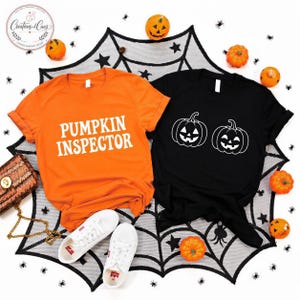 May include: An orange t-shirt with the white text PUMPKIN INSPECTOR and a black t-shirt with a white graphic of two jack-o'-lanterns. The shirts are displayed with small orange pumpkins and a white spiderweb pattern on a white background.