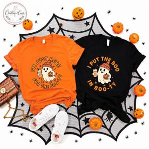 May include: An orange t-shirt with the text I'M JUST HERE FOR THE BOO-TY and a black t-shirt with the text I PUT THE BOO IN BOO-TY. Both shirts feature a graphic of a ghost wearing a beanie, holding a coffee cup and a small jack-o'-lantern.