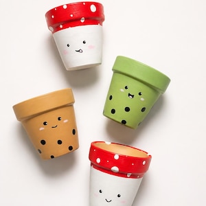 May include: Four small, hand-painted terracotta pots with smiling faces. Two pots have red and white polka dot designs, one is green with black dots, and the last is tan with black dots. The pots are arranged on a white background.