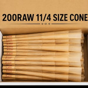 May include: A cardboard box containing 200 light brown paper cones, each marked with the text RAW. The interior lid of the box features the printed text 200RAW 11/4 SIZE CONE. The cones are arranged horizontally in a stack within the open container.
