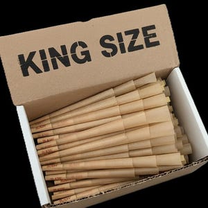 May include: A cardboard box with the text KING SIZE printed in black on the lid, containing a large quantity of light brown, cone-shaped paper rolling papers arranged in a horizontal stack within the open container.