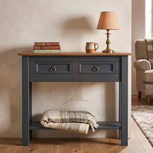May include: A dark grey console table with a natural wood top, two drawers featuring round metal pulls, and a lower shelf. On the table sit two stacked books, a ceramic mug, and a brass lamp with a beige shade. A plaid throw blanket rests on the shelf.