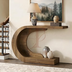 May include: A C-shaped wooden console table with a smooth finish. On top sits a table lamp with a beige shade and a patterned base, plus a small dark wooden box. Two ceramic vases in varying shades of grey and cream rest on the lower shelf.