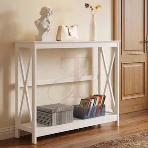 May include: White console table with an X-frame design. The table holds a white bust, a cream-coloured handbag, and a white vase with dried flowers. A striped box and a blue container with books are on the lower shelf.