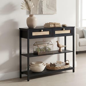May include: A black console table featuring two drawers with light-coloured woven cane panels and small round knobs. The table has two open lower shelves, providing storage space for decorative items, bowls, and a woven basket with linens.