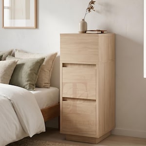 May include: A light oak bedside cabinet with three drawers and a top section. A small vase with dried flowers and a book are on top. The cabinet is beside a bed with pillows and a duvet. The cabinet is approximately 120 cm tall.