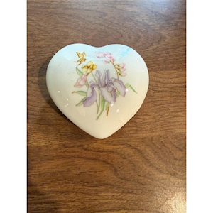 May include: A heart-shaped porcelain trinket box with a smooth cream-colored finish. The lid features a delicate floral illustration of a purple iris, yellow flowers, and pink blossoms with green stems, painted in a soft watercolor style.