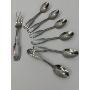 May include: A set of silver-toned stainless steel flatware consisting of one four-tined fork and six spoons. Each piece features a decorative shell motif embossed at the base of the handle, with a smooth, polished finish throughout the set.