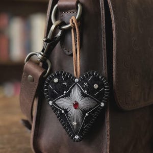 May include: A black heart-shaped felt charm with silver embroidery and a red jewel. The charm is decorated with small crystals and hangs from a leather strap attached to a brown leather bag. The bag has a metal clasp.