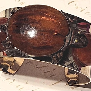 May include: A wide silver-toned cuff bracelet featuring a central dark brown, oval-shaped resin cabochon and two smaller, round, dark brown resin spheres on either side, all set within a dark, textured metal frame shaped like a turtle.