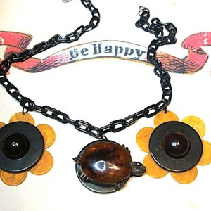 May include: A black chain necklace featuring two flower-shaped pendants with yellow marbled petals and dark centers, and a central turtle-shaped pendant with a brown shell. A pink ribbon graphic with the text be happy is visible.