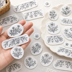 May include: A collection of white fabric patches with black floral embroidery. The patches are in various shapes, including ovals, rectangles, and circles. Some patches feature detailed flower and leaf designs, while others have simple floral borders.