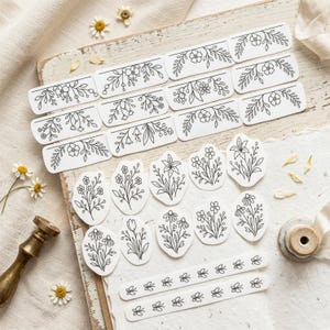 May include: A collection of white stickers featuring black line art illustrations of various floral arrangements, including rectangular border designs, oval-shaped flower bouquets, and two long horizontal strips with repeating flower motifs.