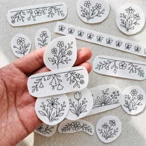 May include: A collection of white felt floral and botanical designs with black line art. The designs include flowers, leaves, and butterflies in various shapes and sizes. Some are rectangular, oval, or circular.