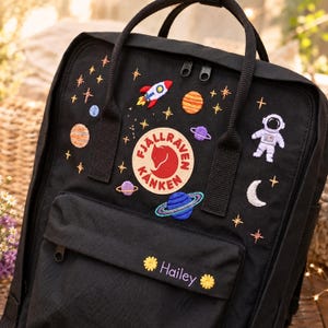 May include: Black backpack with embroidered space-themed patches, including a rocket, planets, stars, and an astronaut. The backpack features a front pocket with the name "Hailey" embroidered in white. The backpack has a top handle and adjustable straps.