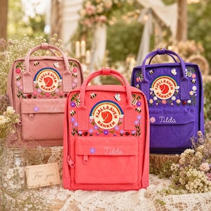 May include: Three Fjällräven Kånken backpacks in pink, coral, and purple, each featuring embroidered flowers, bees, and butterflies. The circular logo patch includes a rainbow, and the front pocket is embroidered with the name Tilda.
