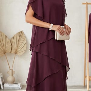 May include: A deep plum-colored midi dress featuring a round neckline, short flutter sleeves, and a layered, tiered skirt design. The dress is paired with a small, rectangular, cream-colored clutch bag and gold-toned heeled sandals with pearl accents.