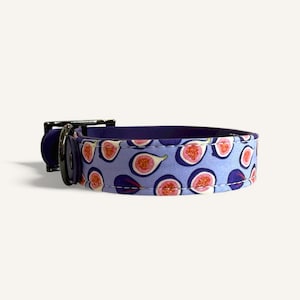 May include: A pet collar featuring a light blue background with a repeating pattern of sliced figs in shades of pink, purple, and white. The collar includes a dark purple buckle and a metal D-ring for attachment, with visible white stitching along the edges.