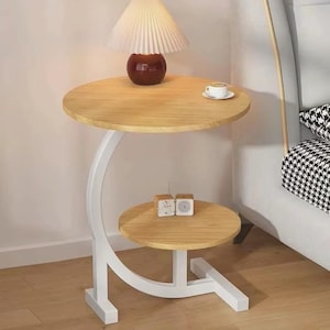 Puede incluir: Una mesa auxiliar de dos niveles con acabado de madera clara y soporte curvo blanco. El nivel superior tiene una lámpara con pantalla color crema y base roja oscura. Una taza y platillo pequeños están en el nivel superior.