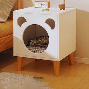 May include: White cat bed with a round opening and two smaller ear-shaped cutouts. A sleeping tabby cat rests inside on a checkered cushion. The bed has wooden legs and a small wooden clock and cat figurine on top.