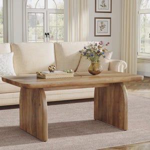 Puede incluir: Una mesa de centro rectangular de madera con acabado natural. La mesa tiene una tapa gruesa y patas curvas. Un sofá beige está en el fondo, junto con un jarrón de flores y objetos decorativos.