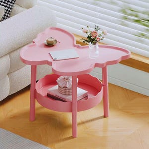 Pode incluir: Uma mesa lateral rosa em forma de borboleta com uma prateleira inferior. A mesa tem um tampo em forma de bandeja com uma xícara pequena e um tablet. Um vaso de flores está sobre a mesa. A prateleira inferior contém um livro e uma caixa decorativa.
