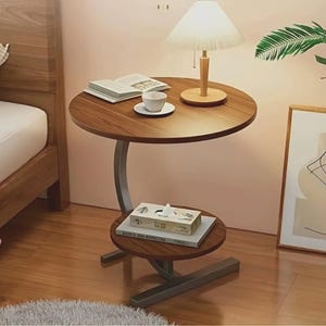 May include: A two-tiered, round side table with a dark wood finish. The top tier holds a lamp, book, and cup. The lower tier has a box and books. The table has a metal frame and stands on a wooden floor.