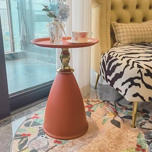 Op de afbeelding: Een decoratieve bijzettafel met een rond, koraalkleurig blad en een bijpassende conische voet. De tafel heeft een goudkleurige, sierlijke centrale steun. Een theekopje en een vaas met bloemen staan op de tafel.