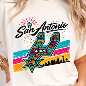 May include: Cream-colored t-shirt featuring a colorful San Antonio Spurs graphic. The design includes the team's logo, the city name, and a skyline silhouette. The shirt has short sleeves and a crew neck.