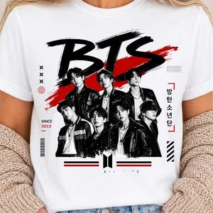 Op de afbeelding: Een wit t-shirt met korte mouwen en een grafische print van de muziekgroep BTS. Het ontwerp bevat de tekst BTS, de woorden SINCE 2013 en Koreaanse tekens. De afbeelding toont zeven leden in zwarte jasjes tegen een rode penseelstreek.