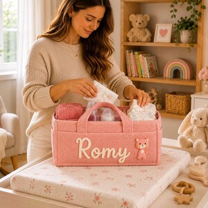 May include: A pink felt baby caddy with the name "Romy" in white script. The caddy contains baby bottles, nappies, and a pink knitted item. A small teddy bear is attached. The caddy is on a changing table.