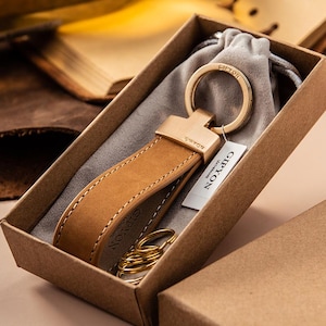 May include: A tan leather keychain with a gold-coloured ring and hardware, presented in a brown cardboard box. The keychain has a stitched design and the brand name "GIPYON" is visible on the tag and the leather. A small grey pouch and gold keyrings are also in the box.