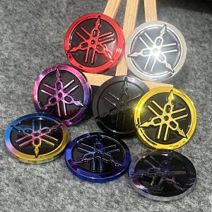 Op de afbeelding: Een verzameling ronde, metalen emblemen met het Yamaha-logo in verschillende kleuren. De emblemen bevatten rode, zilveren, zwarte, gouden, blauwe en iriserende afwerkingen. Het Yamaha-logo is een gestileerd, drietandig ontwerp.
