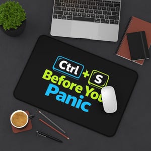 May include: Black rectangular mousepad with the text "Ctrl + S Before You Panic" in blue and green. A white computer mouse is on the right side of the mousepad. A laptop, coffee cup, and notebooks are also visible.