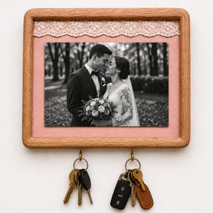 Peut inclure: Un porte-clés mural en bois rectangulaire présentant une photographie en noir et blanc d'un couple en tenue de mariage. Le cadre est agrémenté d'un fond en tissu rose clair et d'une bordure en dentelle blanche, avec deux crochets en laiton.