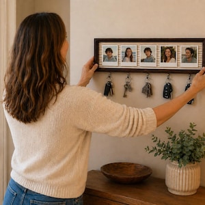 Peut inclure: Un porte-clés mural rectangulaire en bois foncé doté d'une rangée horizontale de cinq cadres photo à bordure blanche. Sous les cadres se trouvent cinq crochets métalliques, chacun portant un trousseau de clés. Le cadre a un fond rayé.