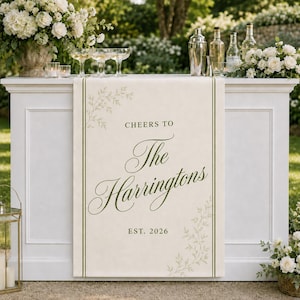 May include: A cream-colored fabric table runner featuring a dark green border and botanical leaf illustrations. The text reads: CHEERS TO The Harringtons EST. 2026. The runner is draped over a white bar, positioned below glassware and floral arrangements.