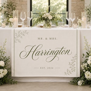 May include: A white fabric table runner featuring the text MR. & MRS. Harrington EST. 2026 in dark green lettering. The design includes delicate green leaf illustrations in the top left and bottom right corners, framed by thin vertical lines.