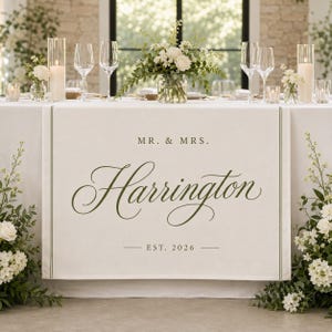 May include: A white fabric table runner featuring the dark green printed text MR. & MRS. Harrington EST. 2026. The runner has thin vertical green lines along the sides and is draped over a table set with white flowers, candles, and glassware.