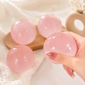 Peut inclure: Trois sphères en quartz rose polies et lisses, présentant une teinte rose pâle translucide. Les sphères ont une forme ronde avec une surface brillante et réfléchissante. Une sphère est tenue au premier plan, tandis que deux autres reposent sur un plateau en bois.
