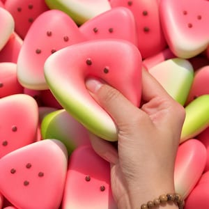 Könnte beinhalten: Eine Hand drückt einen wassermelonenförmigen Stressball zusammen. Der Stressball ist rosa mit braunen Samendetails und einer grünen Schale. Viele ähnliche Stressbälle befinden sich im Hintergrund.