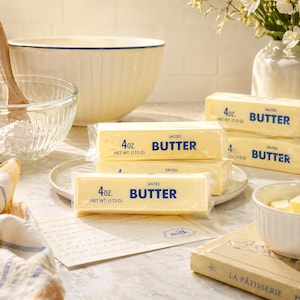 May include: A selection of salted butter sticks, each weighing 113 g, are displayed. A large mixing bowl, a glass bowl with flour, and a small white bowl with butter cubes are also present. A recipe card and a vase of flowers complete the scene.