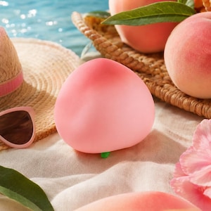 Peut inclure: Gros plan sur un objet en forme de pêche de couleur rose clair, posé sur une surface sablonneuse. Un chapeau de paille et des lunettes de soleil sont visibles en arrière-plan, ainsi que des pêches fraîches et un panier tressé. L'ensemble évoque l'été.