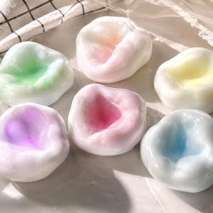 Könnte beinhalten: Sechs abgerundete, glänzende, halbtransparente Objekte mit einer glatten, gewölbten Oberfläche. Jedes Stück hat einen Kern in einer anderen Pastellfarbe, darunter Grün, Rosa, Gelb, Violett, Magenta und Blau, eingebettet in milchig-weißes Material.