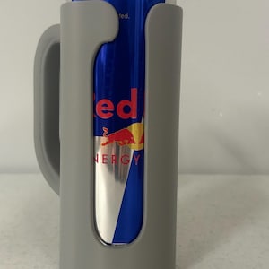May include: A gray can cooler with a handle, holding a blue and silver Red Bull energy drink can. The can has the word "Red Bull" in red and a yellow and red logo.