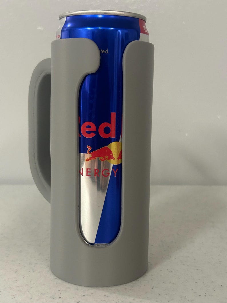 May include: A gray can cooler with a handle, holding a blue and silver Red Bull energy drink can. The can has the word "Red Bull" in red and a yellow and red logo.