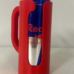 May include: A red can cooler with a handle holds a blue and silver Red Bull energy drink can. The cooler has a curved design to fit the can snugly. The Red Bull can has the word "Red" in large letters.